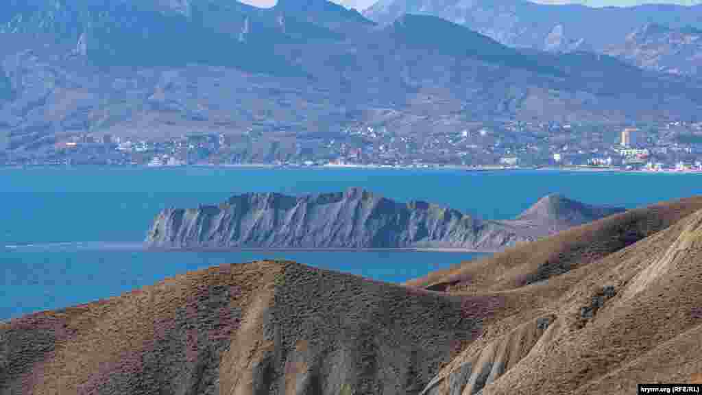 Мыс Хамелеон на фоне поселка Коктебель. Свое название Хамелеон получил из-за постоянно меняющегося окраса и причудливой формы. Цвет мыса Хамелеон напрямую зависит от времени суток и погодных условий. В пасмурную погоду он синий или голубой, в солнечную – от желтого до светло-красного