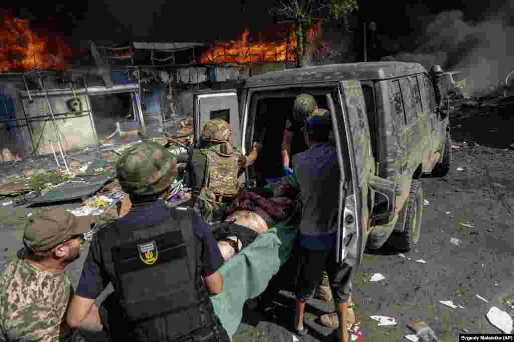 Пораненого чоловіка несуть у машину швидкої допомоги, на задньому плані палає пожежа