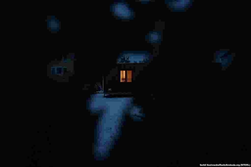 Крізь отвори у посіченому обстрілами паркані видно світло у вікні модульного будиночка і згорілий автомобіль на подвір'ї