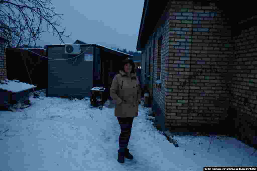 Жителька Мощуна Любов Тополь стоїть між модульним житлом, яке їй надав благодійний фонд, і своїм згорілим будинком. Вона втратила на війні єдиного сина Олександра, який був командиром місцевої тероборони.