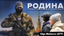 Люди проходят мимо стенда с изображением российского военнослужащего и надписью "Родину, которую мы защищаем" на уличной выставке плакатов на военную тематику в центре Санкт-Петербурга