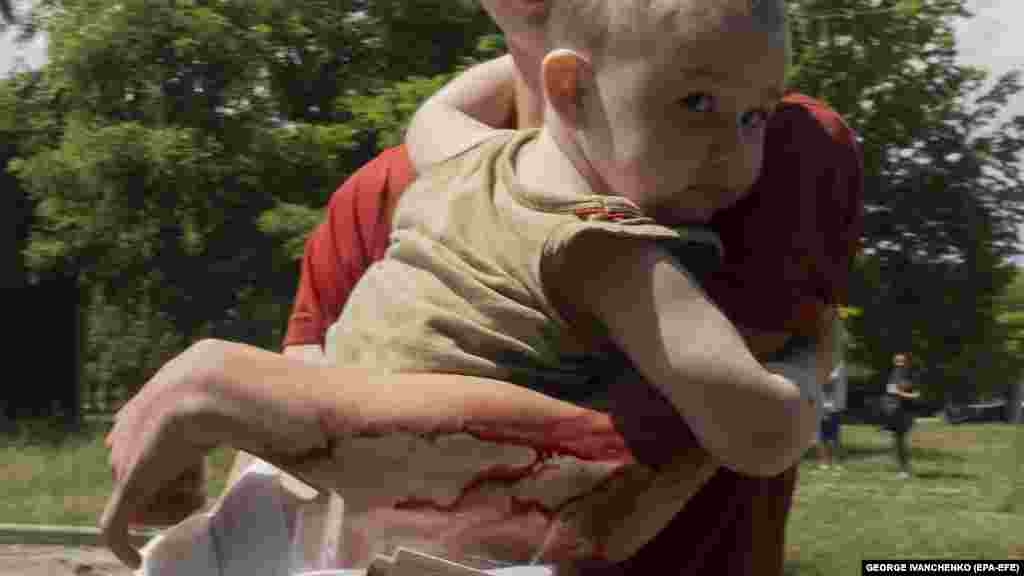 Травмований підліток несе на руках малюка, покидаючи місце ракетного удару у Первомайському. 4 липня 2023 року Російська армія від моменту широкомасштабного вторгнення в Україну систематично піддає ракетним, артилерійським обстрілам і дроновим атакам українські цивільні об'єкти, зокрема житлові будинки, лікарні і школи.