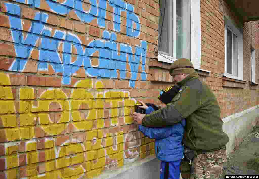 Чоловік і хлопчик розмальовують кольорами українського прапора напис на стіні в Куп’янську: «Любіть Україну. Любіть Шевченка»