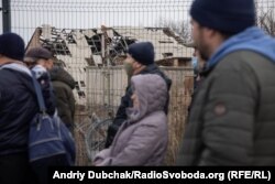 Люди стоять у черзі до модуля прикордонників на тлі зруйнованих у 2014-2015 роках будинків