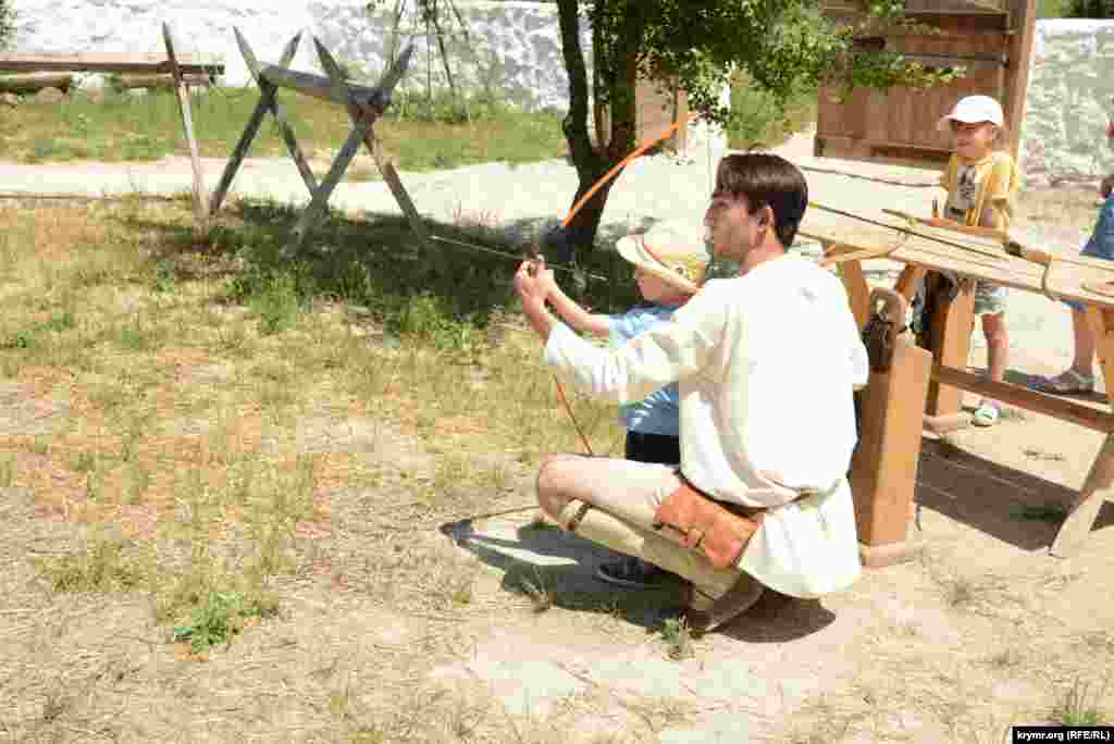 Реконструктор помогает мальчику-посетителю стрелять из лука по мишени 