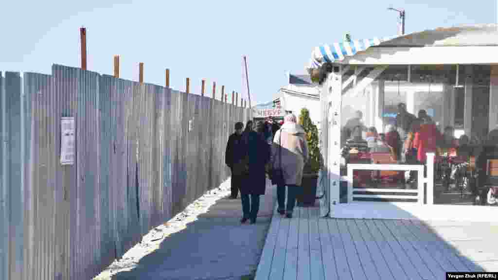 Проход к морю через узкий коридор между строительным забором и торговыми павильонами 