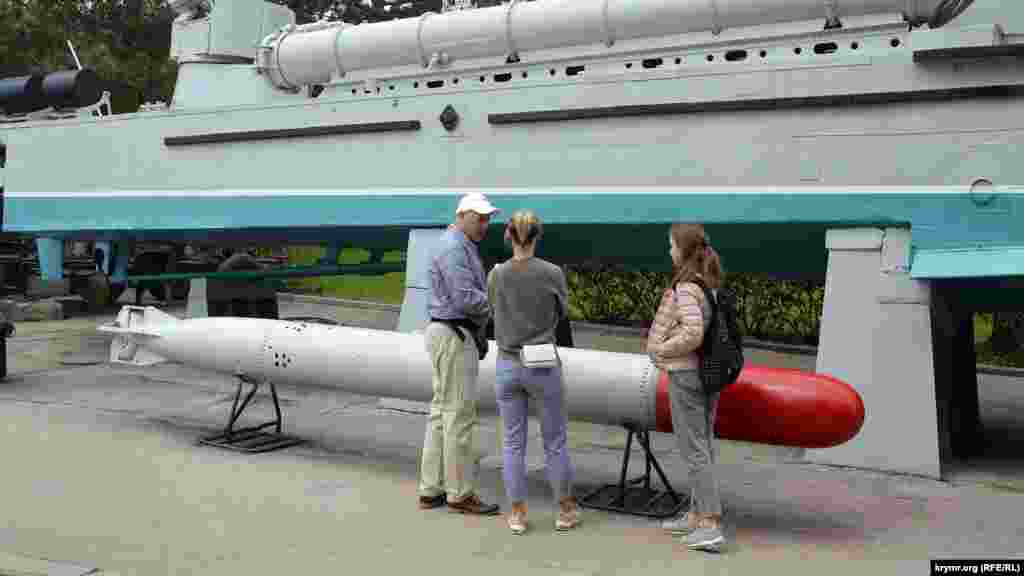 Посетители рассматривают находящуюся под открытым небом военную технику и вооружение – бронекатер и торпеду