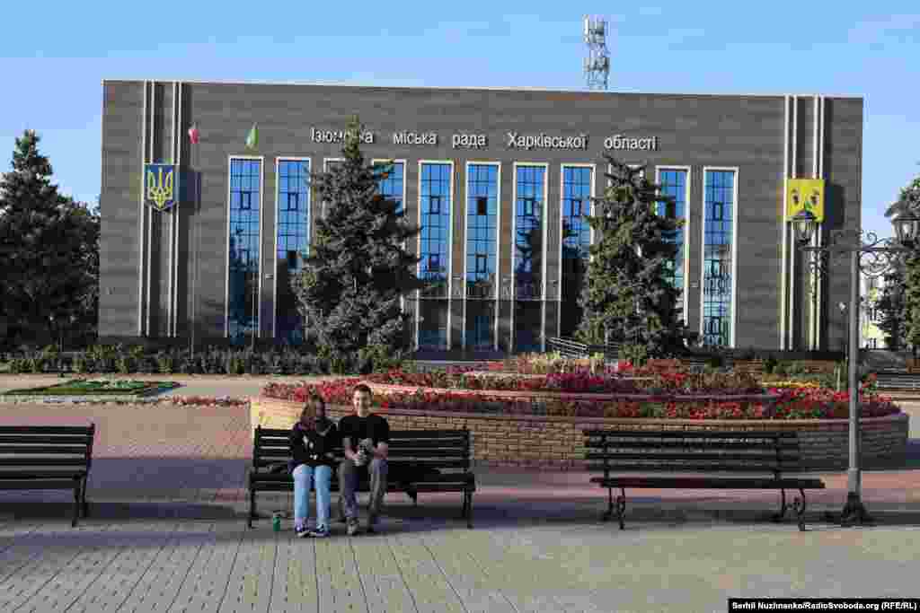 Жителі Ізюму в центрі міста. Не на камеру багато хто з місцевих жителів розповідає про окупацію, голодні смерті і як за їжу розбирали завали під керівництвом російських військових. Позаду спалена  будівля місцевої міськради, вересень 2022  року / Праворуч: початок жовтня 2024-го. Більшість нинішніх мешканців Ізюму – це люди пенсійного віку. Молоді й активні повертаються неохоче. За словами місцевих жителів, в Ізюмі дуже повільно йде відновлення пошкодженого житла. Через це будь-які інші ремонти, що відбуваються в місті, наприклад, будівлі мерії в центрі, що здійснюється в межах цільової міжнародної програми, викликають невдоволення в місцевих. Адже навколо побите й обгоріле житло, початок жовтня 2024 року