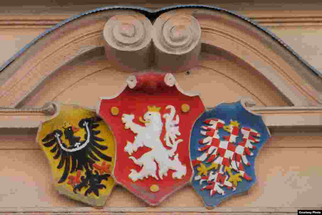 Это сложный герб земель Чешской короны над воротами обычного жилого дома на Ржегоржовой улице. Ладислав Ржегорж - еще один чешский герой, только другой эпохи: почтовый чиновник и активист военно-патриотического клуба "Сокол", он в 1942 году был казнен немецкими оккупантами. На центральном гербовом щите - двухвостный белый лев (Чешские земли), слева - черный орел в золотом поле (Силезия), справа - красно-белый клетчатый орел на синем поле (Моравия).