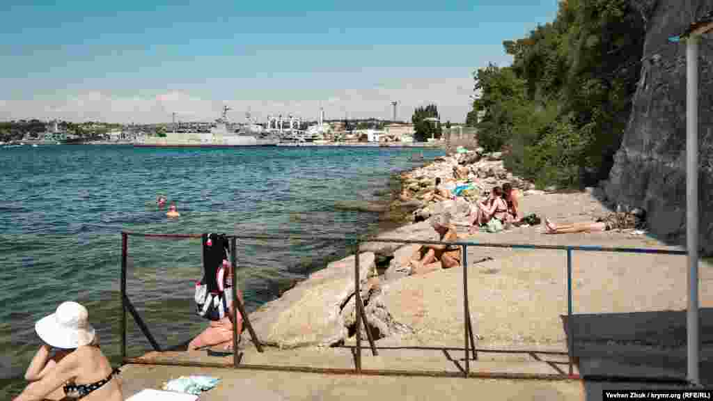 За огорожею офіційного пляжу – невеликий дикий пляж, на якому серед каменів теж засмагають люди. На дальньому плані видно російські військові кораблі