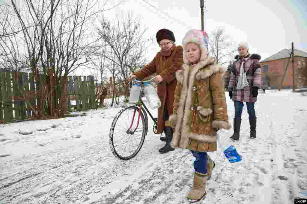 Ніна (ліворуч) та її онуки Діана, 14 років (праворуч), і 6-річна Саша (у центрі) ідуть набрати води з криниці, що розташована на околиці села. Ніна пояснює: «На початку вулиці є водопровід, але часто води не вистачає. Ми набирали воду, але тиск у трубі став занадто слабким, тому ми перестали. Набираємо воду зі свердловини та криниці. Але насос зламався, тому зараз можемо скористатися лише криницею». Будинок цієї сім’ї розташований на відстані 15 кілометрів від лінії розмежування в Торецьку Донецької області. Сестри Діана і Саша живуть зі своєю бабусею в цьому будинку з початку конфлікту у 2014 році.