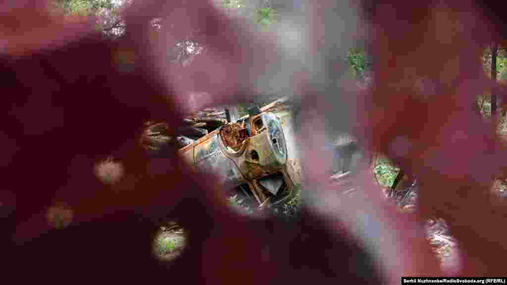 Крізь отвори у посіченому обстрілами паркані видно згорілий автомобіль на подвір'ї одного з будинків