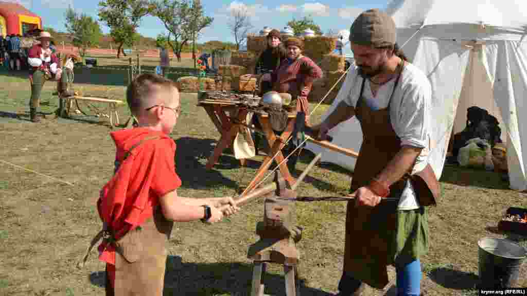Маленький посетитель и реконструктор-викинг выковывают детский нож на наковальне 