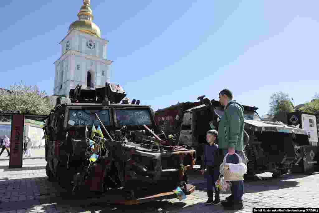 На площі біля храму експонується розбита російська військова техніка. Батько та син несуть великодній кошик і розглядають її. Київ, 20 квітня 2025 року