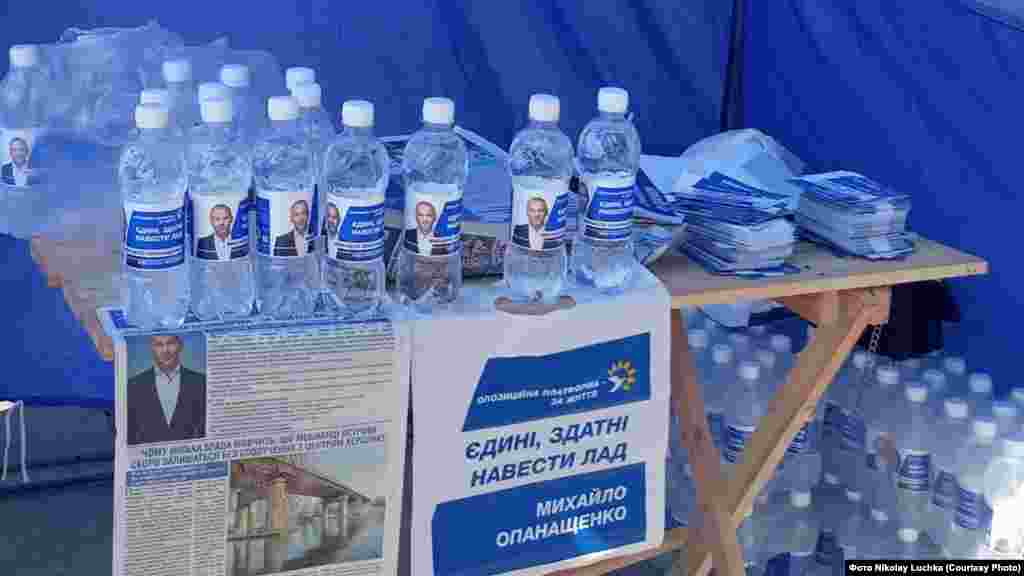 Де? Херсон. Що роздають? Воду. Від кого? Михайло Опанащенко, потенційний кандидат в мери.  Деталі: Пляшки з водою, фото Опанащенка, з логотипом і гаслом ОПЗЖ почали безкоштовно роздавати у брендованих наметах партії і громадському транспорті Херсону ще до офіційного старту кампанії. Воду для цього набирали просто з системи централізованого водопостачання. Сам Опанащенко назвав це «турботою про людей»: «Якщо ви вважаєте, що хтось проголосує за пляшку води, ви помиляєтеся&raq