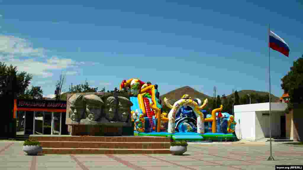 Монумент «Воинам-участникам героического десанта, павшим за крымскую землю в годы Великой Отечественной войны» в окружении батутов и других аттракционов. Площадь украшает флагшток с российским стягом